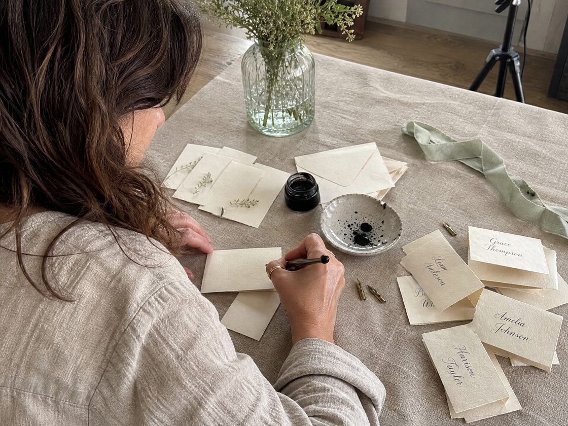 The maker at her linen-covered table, seen from behind, lettering a place card with a pointed pen. Finished cards with guests&rsquo; names, spare nibs, a jar of black ink and a sage silk ribbon are spread around her.