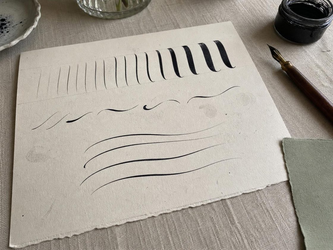 A practice sheet on a linen table showing rows of thin and thick pressure strokes in black ink, with a pointed pen and ink jar beside it.
